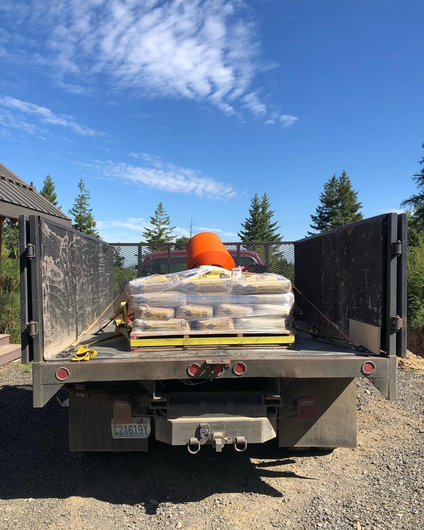 Back of a flatbed truck carrying pallets of concrete mix, an orange bucket, and a yellow safety strap, with trees and a blue sky in the background.