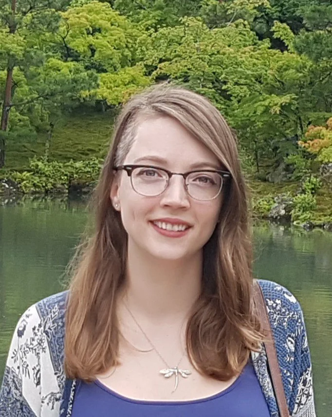 A young woman with glasses and shoulder-length brown hair, smiling, standing outdoors near a body of water with lush green trees in the background.