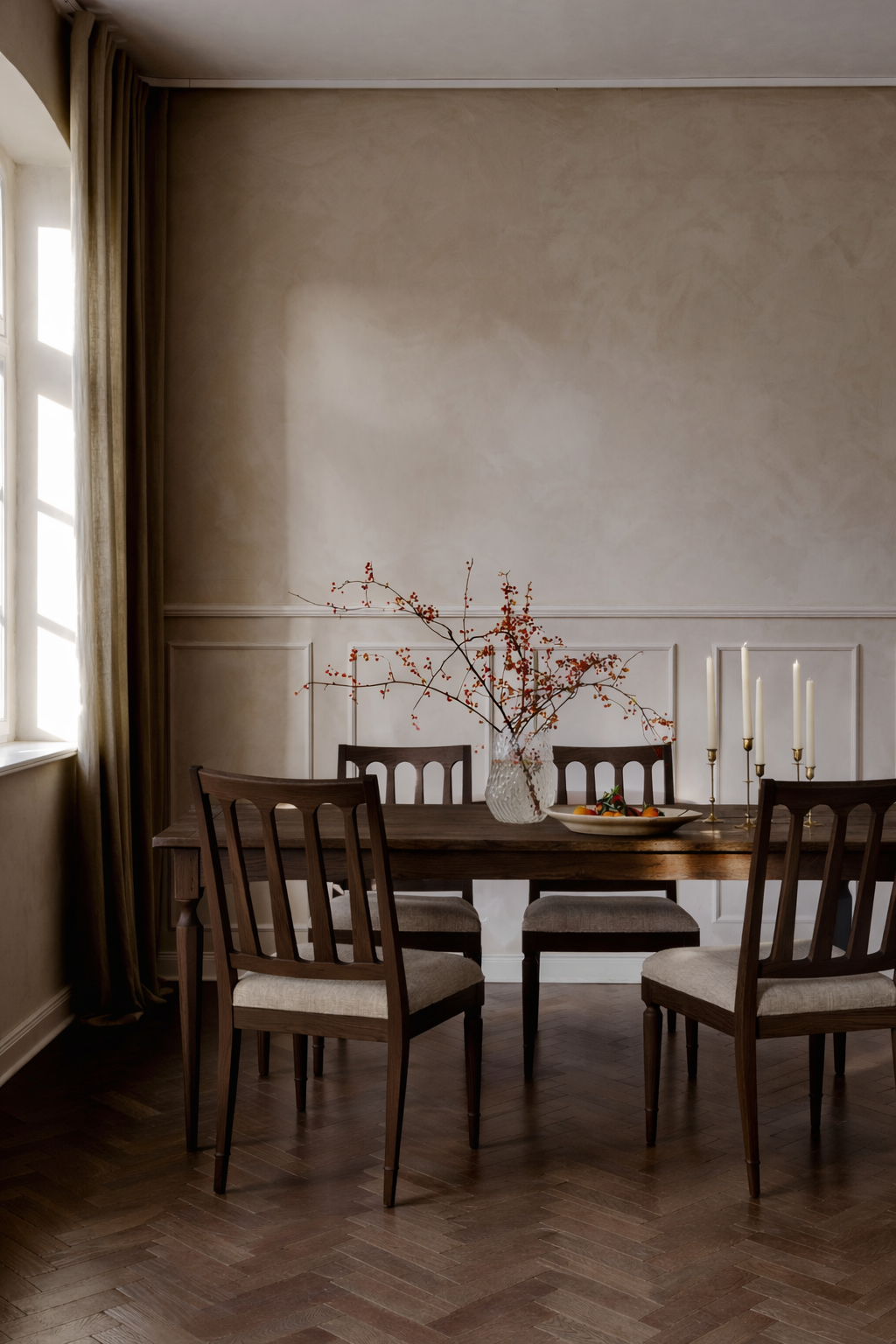 A wooden dining table with five chairs, a glass vase with branches and red berries, a bowl of fruit, and brass candlestick holders with white candles is set near a window with curtains in a warmly lit room.
