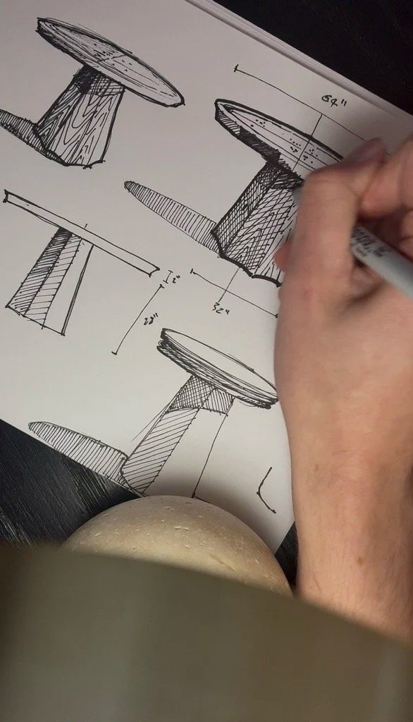 Hand sketching design plans for a small round table with a thick, textured pedestal base on a sheet of paper.