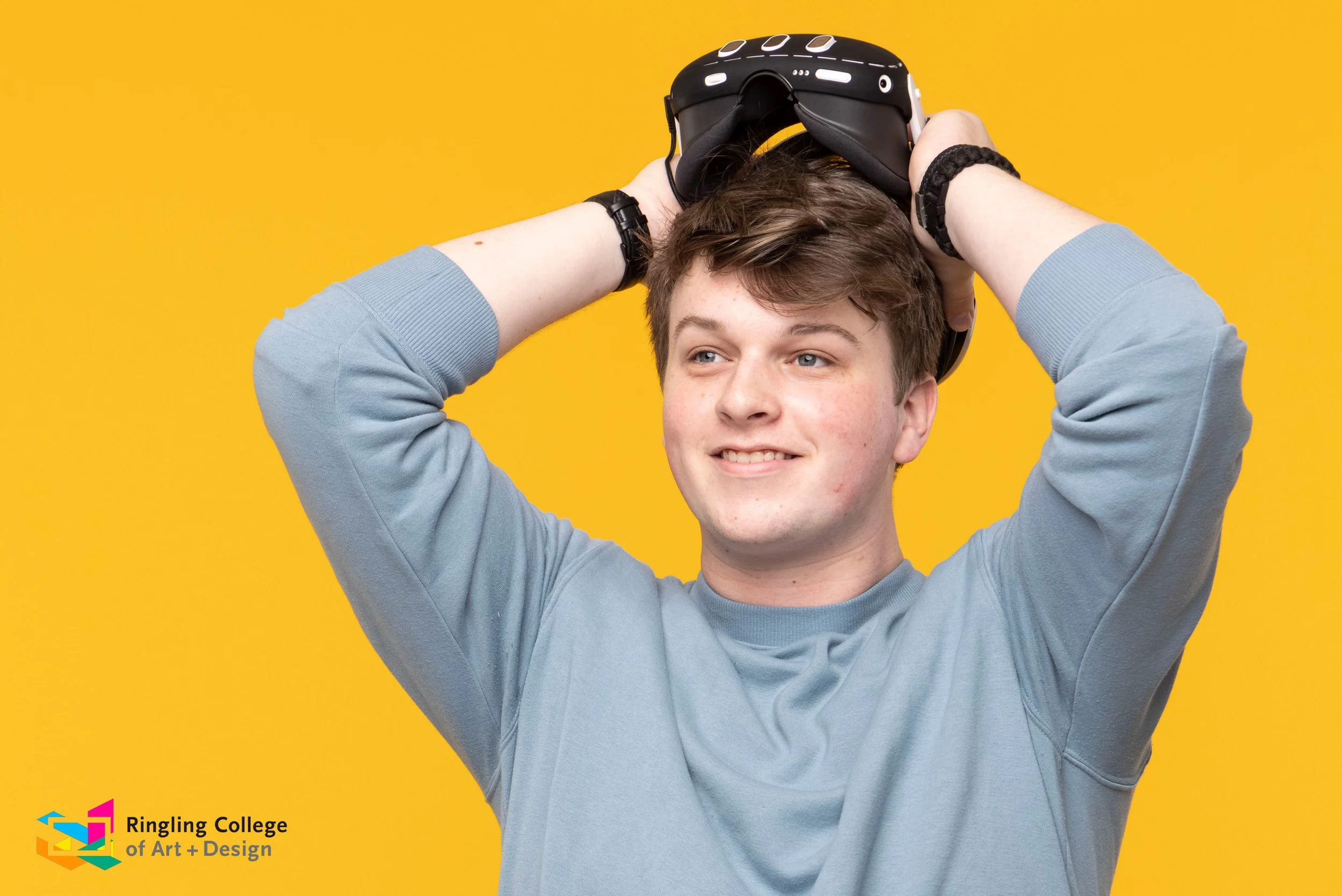A young man with short brown hair and a light grey sweatshirt is smiling and holding a virtual reality headset above his head against a yellow background. The logo for Ringling College of Art + Design is in the bottom left corner. Eli McBride