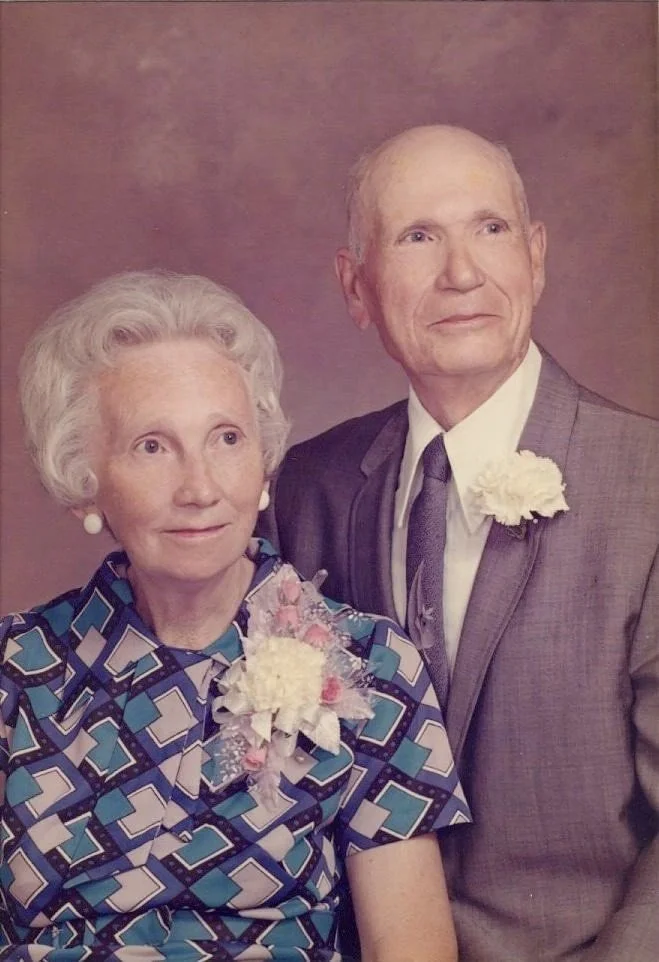An elderly woman and man dressed in formal attire, with corsages on their clothing, posing together for a portrait against a purple backdrop.