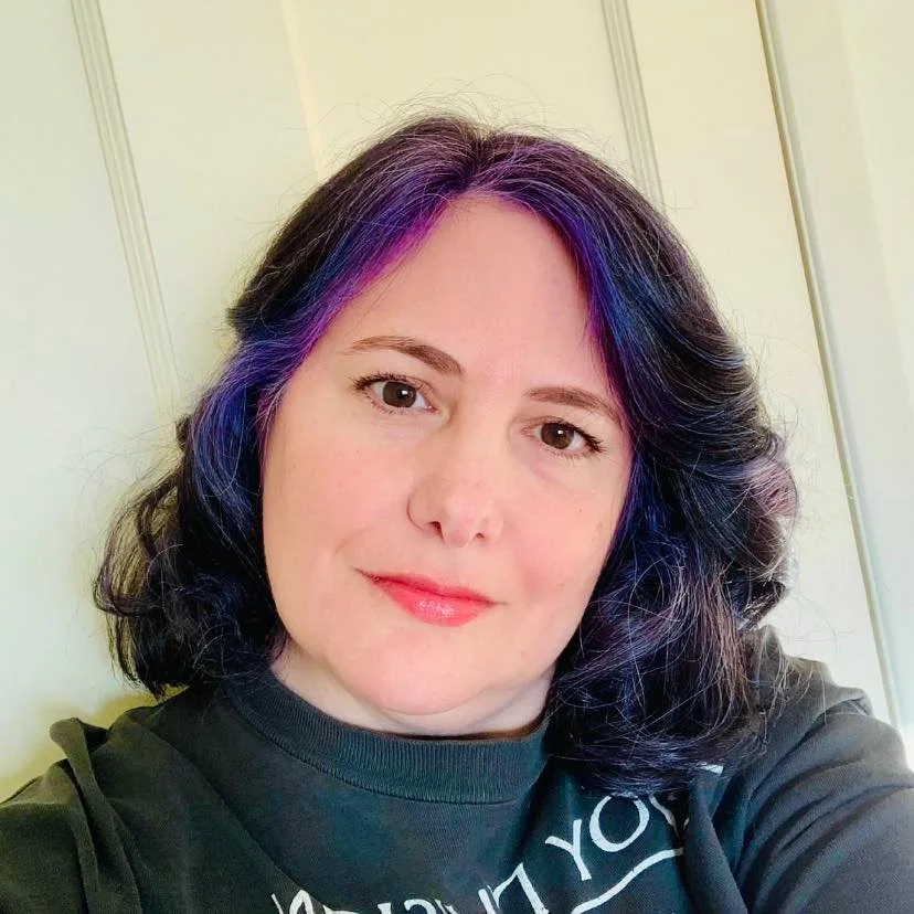A woman with shoulder-length dark hair with purple and blue streaks, smiling, wearing a black shirt, standing in front of a light-colored wall.