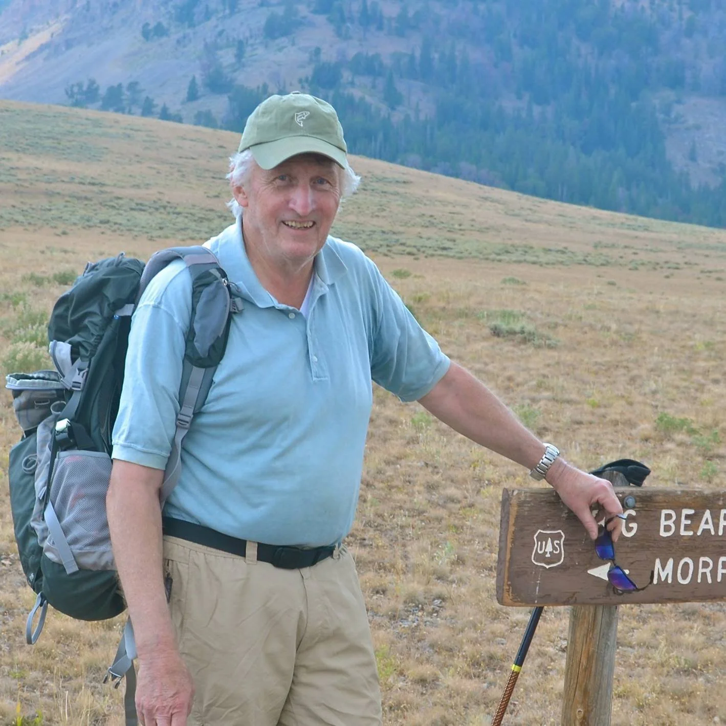 Older man with a backpack and a green cap standing next to a wooden sign in a grassy mountainous area.