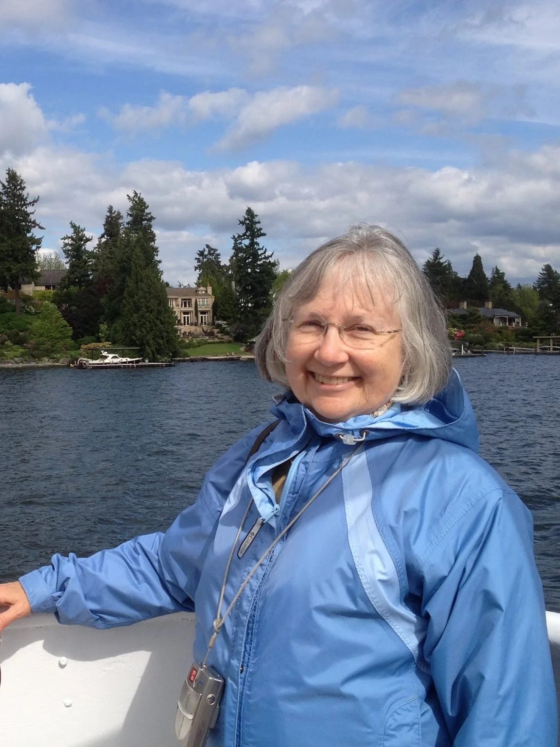 An elderly woman with gray hair and glasses smiling, wearing a blue jacket, standing on a boat with water and houses on the shoreline behind her under a partly cloudy sky.