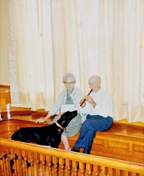 An elderly woman and man sitting on a wooden stage with a curtain behind them. The woman is holding a dog, and the man is playing a recorder. The dog is lying on the woman's lap.
