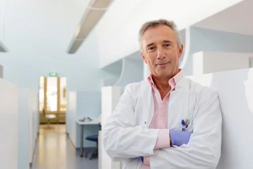 A confident middle-aged male doctor standing in a modern hospital corridor with his arms crossed, wearing a white lab coat and a pink shirt.