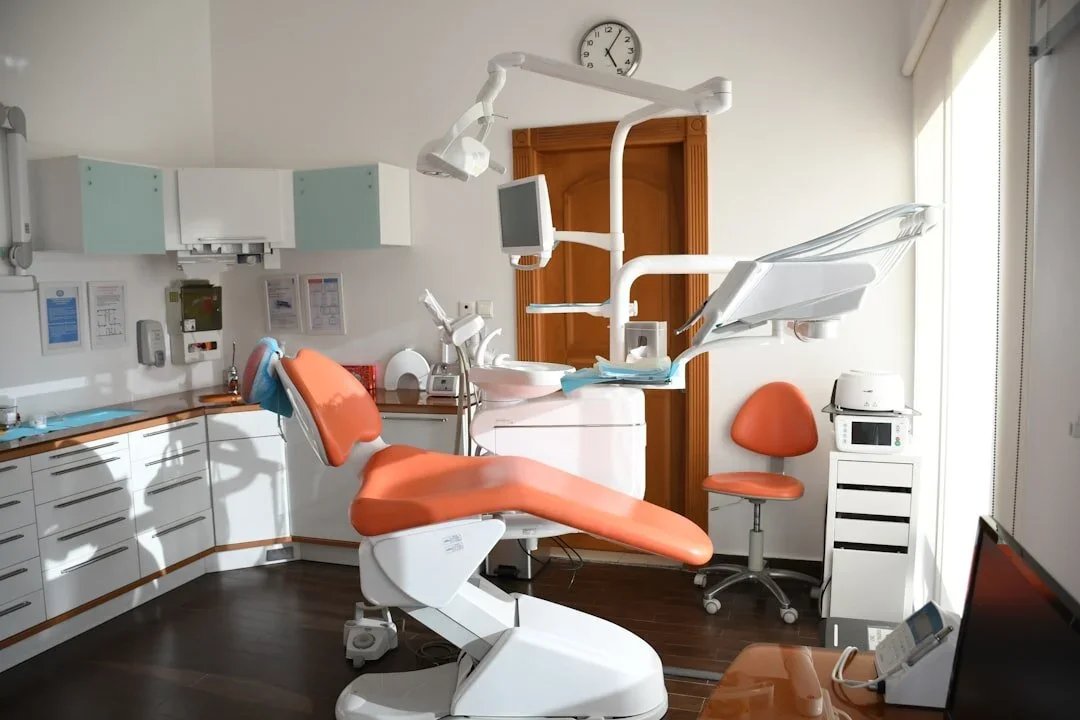 Empty dental examination room with orange dental chair, dental equipment, and a window with blinds.