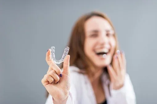 Woman holding a small orthodontic retainer in front of her face, smiling and laughing.