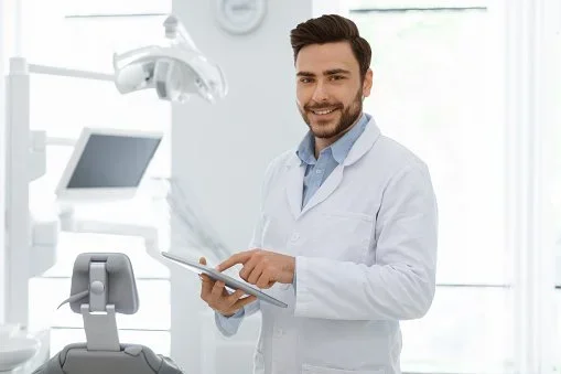 A male doctor with a beard and dark hair, wearing a white lab coat, is smiling and holding a tablet in a modern dental clinic.