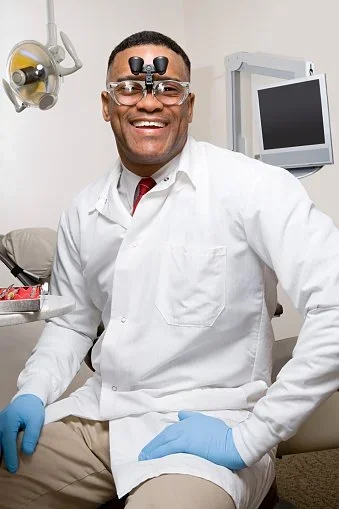 A smiling male dentist in a dental office, wearing a white coat and blue gloves, with dental equipment and a monitor in the background.