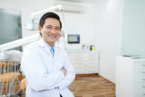 A smiling male dentist in a white coat standing in a dental office with dental equipment and cabinetry in the background.