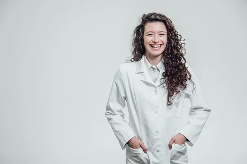 Young woman with curly hair smiling, wearing a white lab coat, standing against a plain light background.