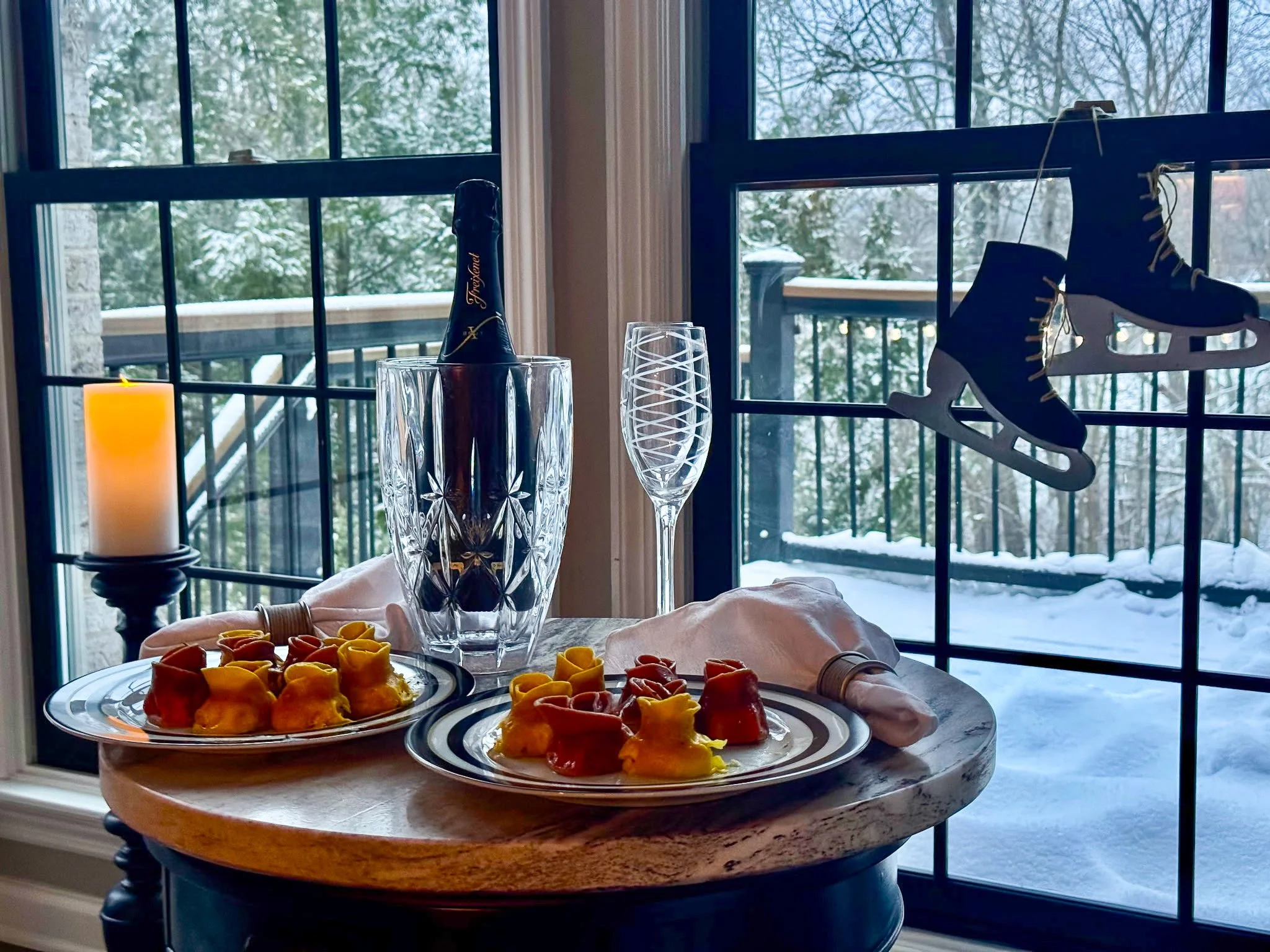 Holiday table set with champagne, plates of tortellini, a lit candle, and ice skates hanging in front of a snowy window.