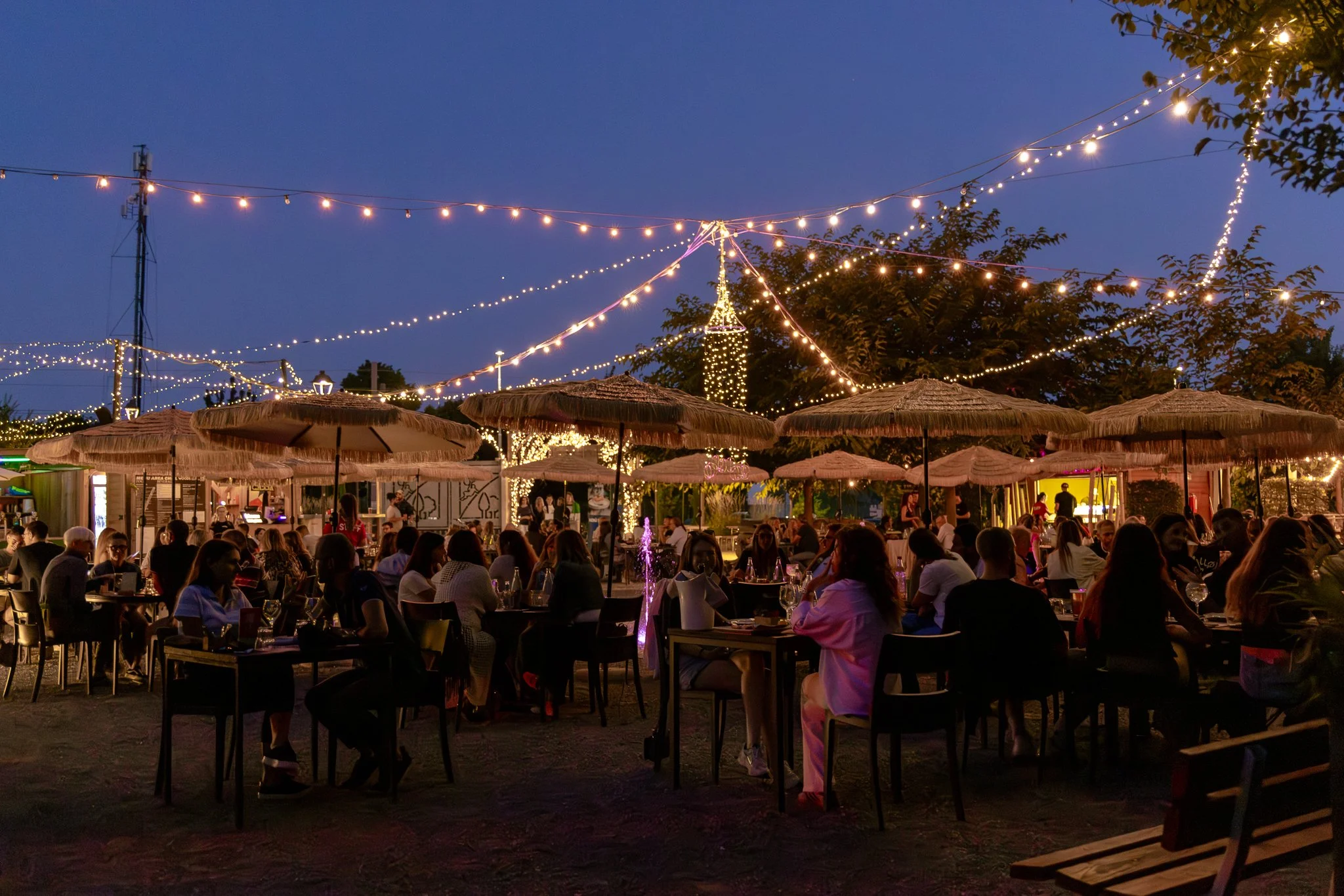 Una scena di una festa all'aperto di sera con molte persone sedute ai tavoli sotto ombrelloni di paglia e decorazioni luminose sospese nel cielo blu.