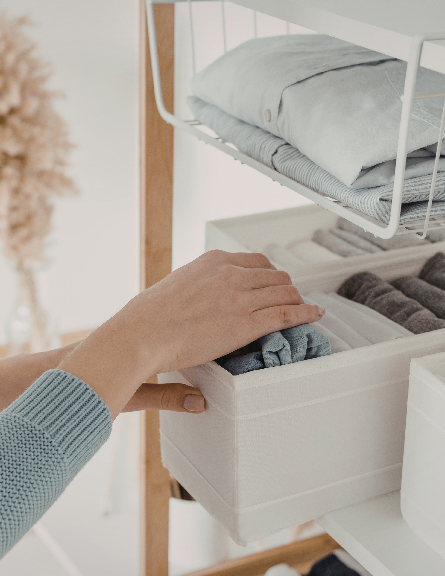 Person placing folded clothes into a white storage box on a shelf. The shelf has multiple compartments filled with neatly folded clothing and textiles in neutral shades.