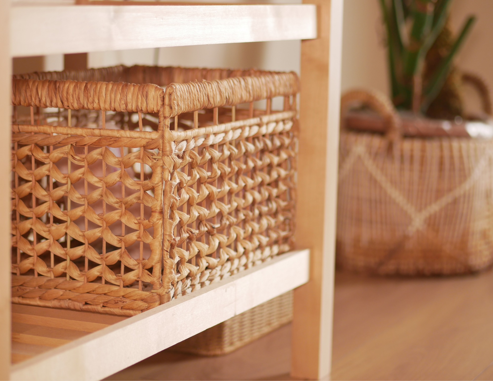 Close-up of a woven basket with a basket pattern, placed on a wooden shelf. In the background, blurred baskets on another shelf with a plant on the right side.