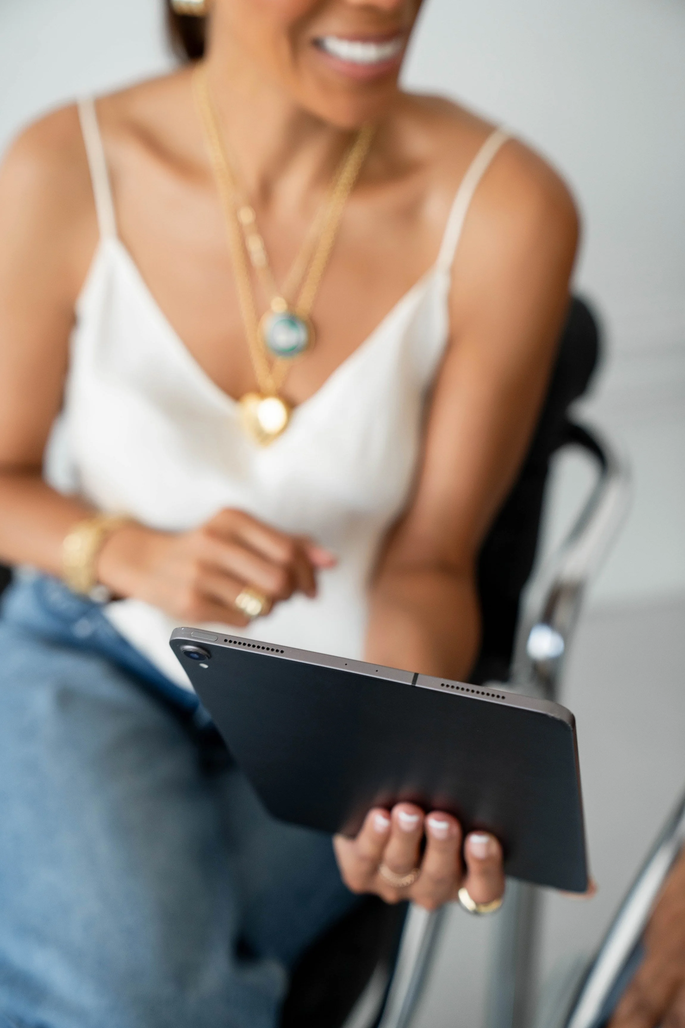 A woman in a white spaghetti strap top is sitting at a table, holding a smartphone in her left hand and appearing to look at it. She is smiling and wearing layered gold necklaces and rings. The background shows part of an office chair and a table.