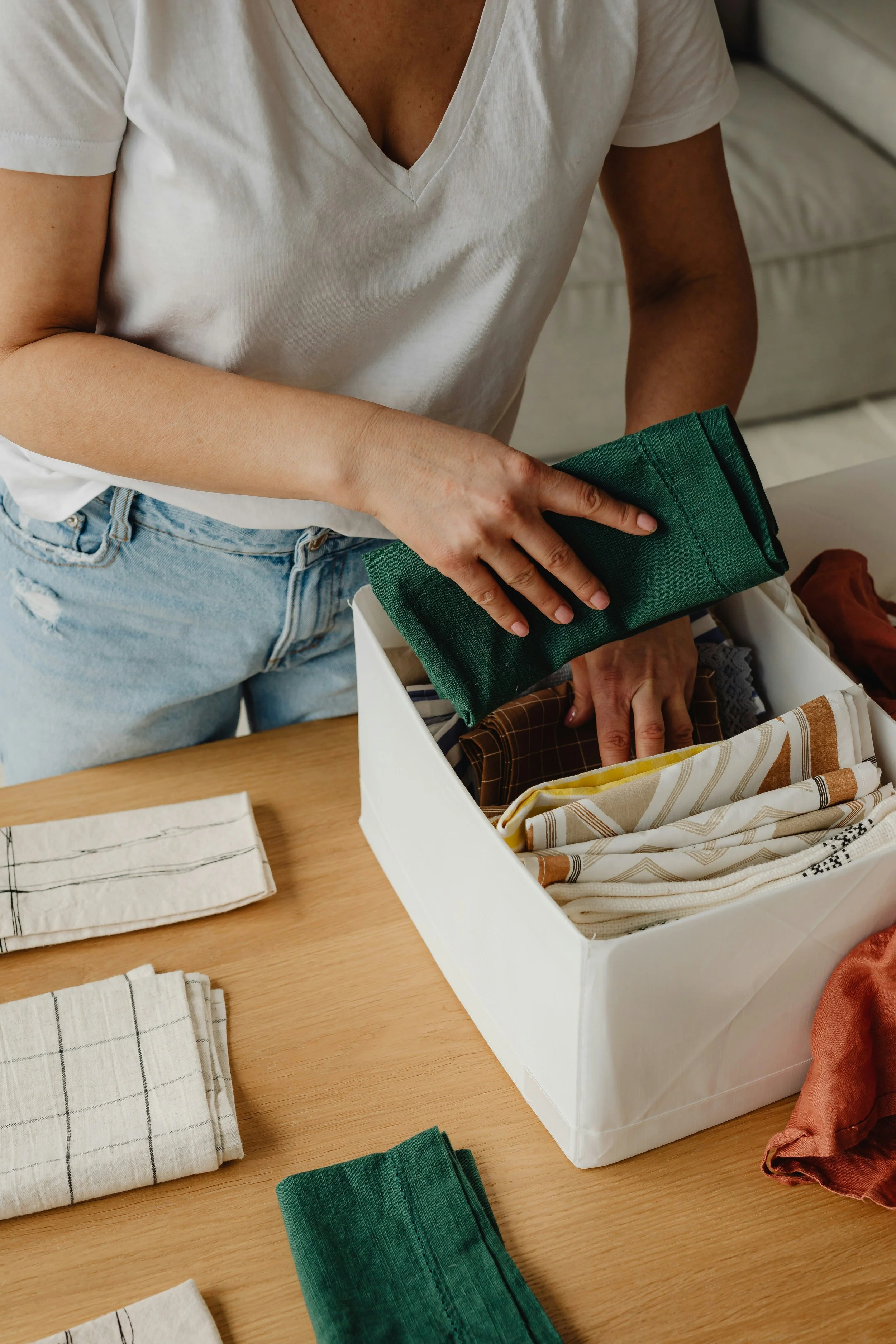 A person is folding or sorting fabric scraps on a wooden surface, with various fabric pieces and a white storage box filled with fabric pieces around them.