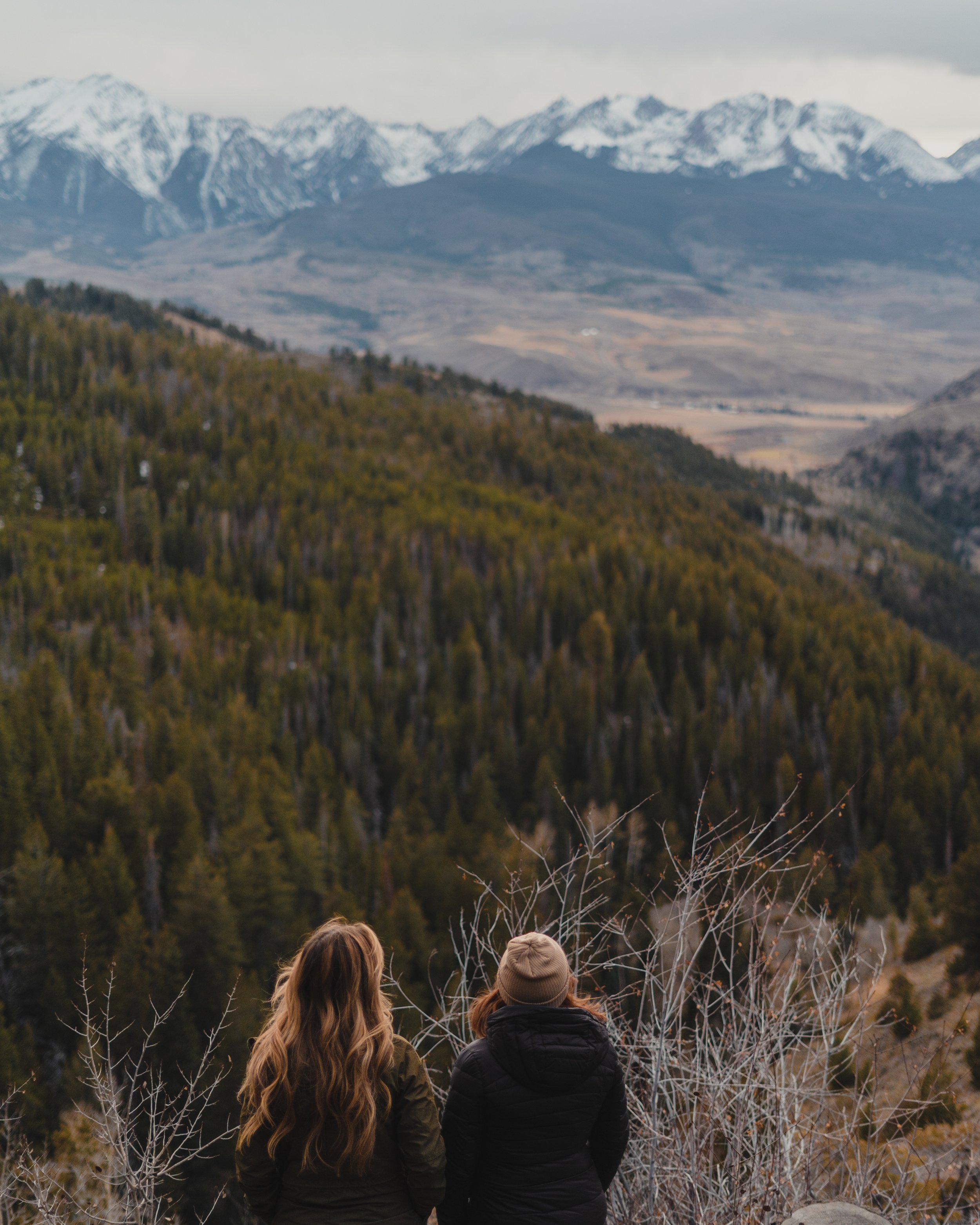 Two women with long hair, wearing jackets, standing on a hill overlooking a forested valley with mountains in the distance, some snow-capped.