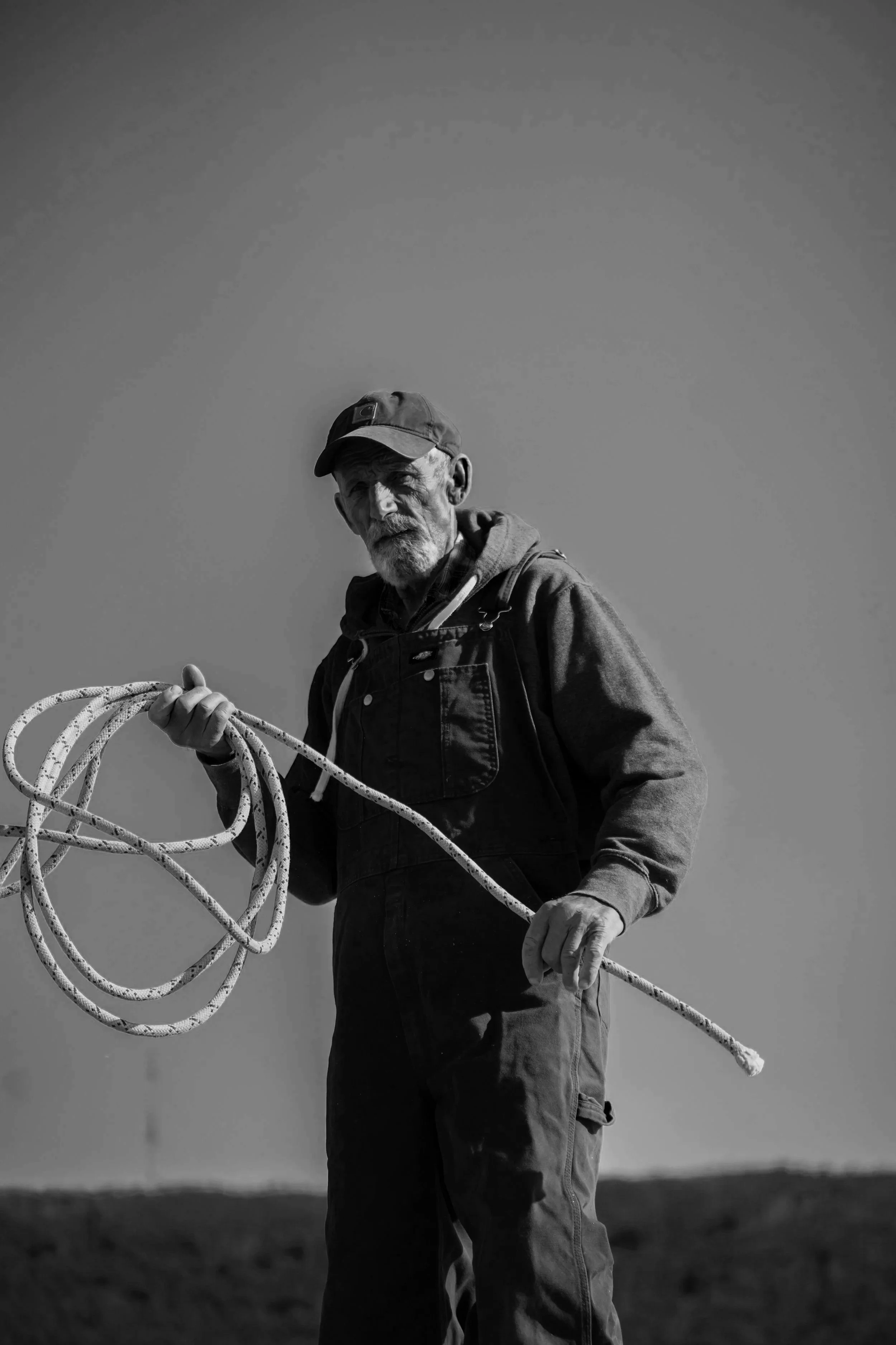 An elderly man holding a coiled rope outdoors, wearing a cap, hoodie, and overalls, with a plain background.