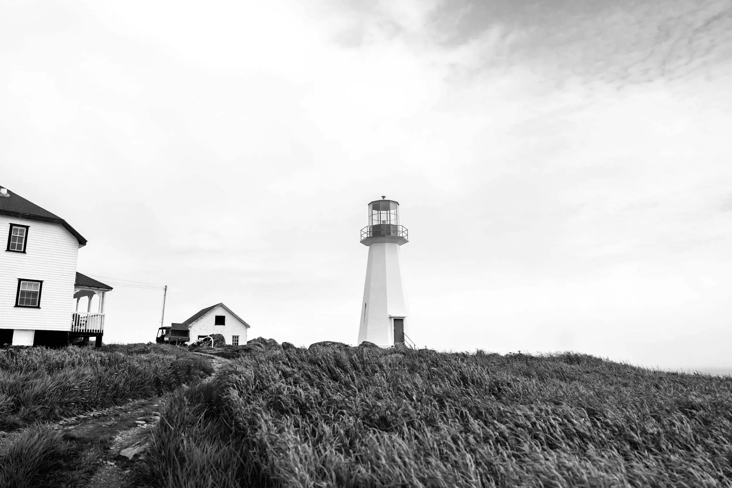 Black and white photo of a lighthouse on a grassy hill next to a house and small building, with a cloudy sky in the background.