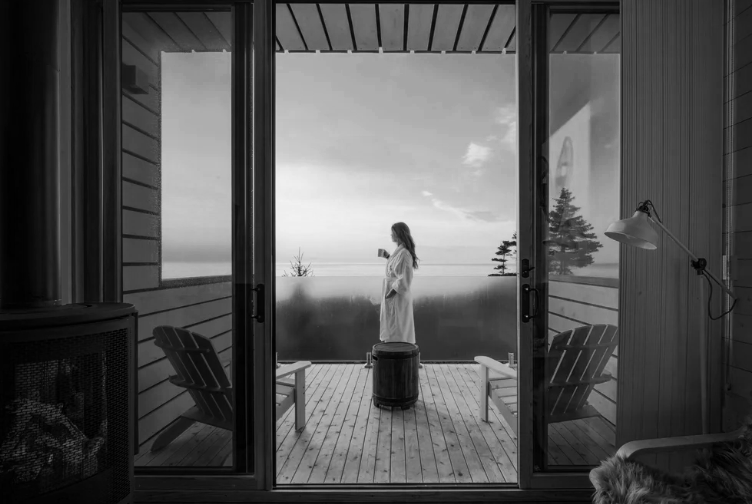 A woman in a robe standing on a balcony, looking at her phone, with two outdoor chairs and a small table, overlooking a foggy landscape with trees.