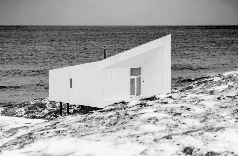 A modern white house built on stilts on a rocky beach, overlooking the ocean.