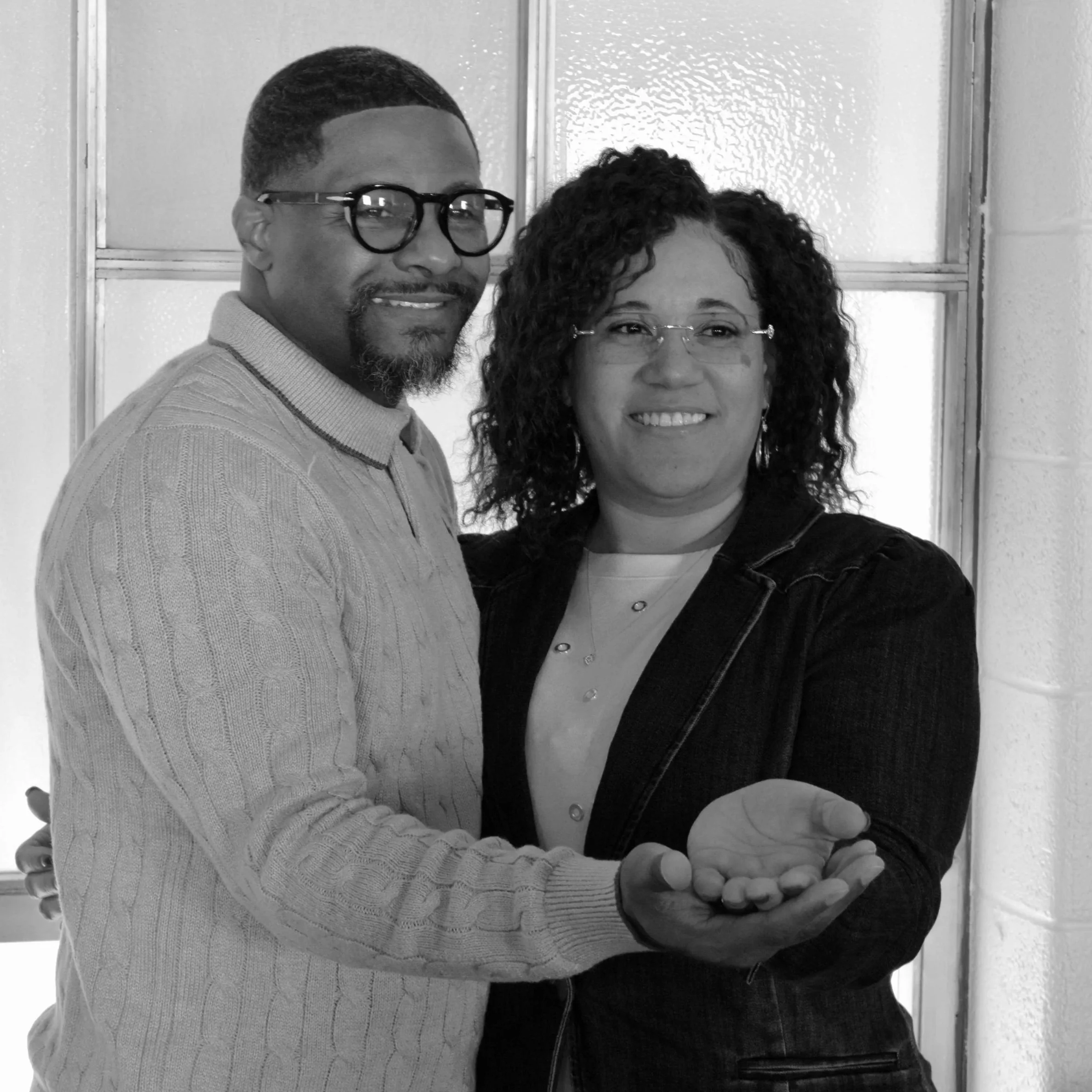 A man and a woman are standing close together, smiling at the camera. The man is wearing glasses and a light-colored, textured sweater. The woman has curly hair, glasses, and is wearing a dark top with a light shirt underneath. They are indoors, in front of a wall with a window or glass panel.