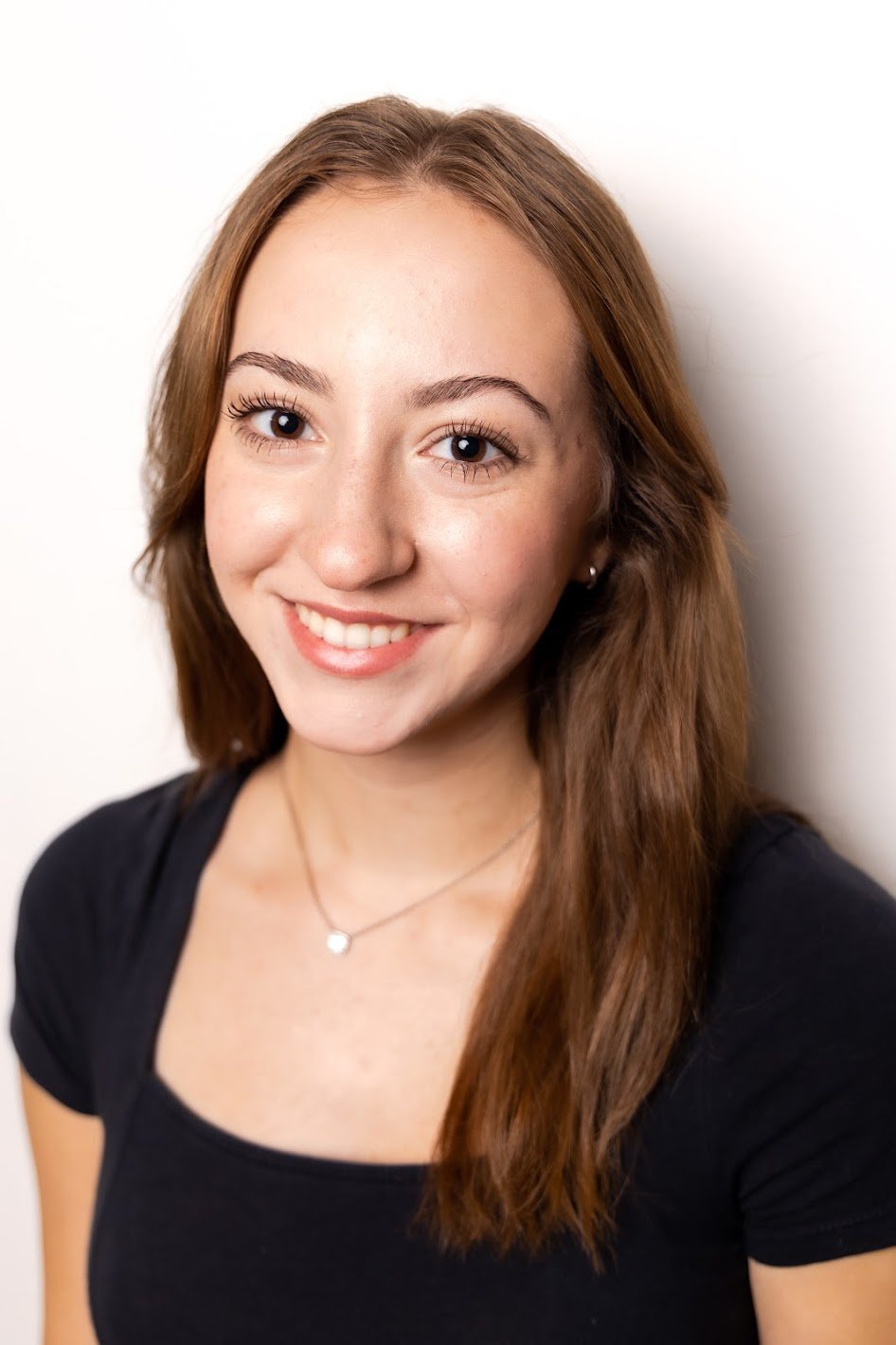 Close-up of Jiana Casazza, a young actress with long, brown hair, smiling and wearing a black top, small earrings, and a delicate necklace, standing against a white wall.