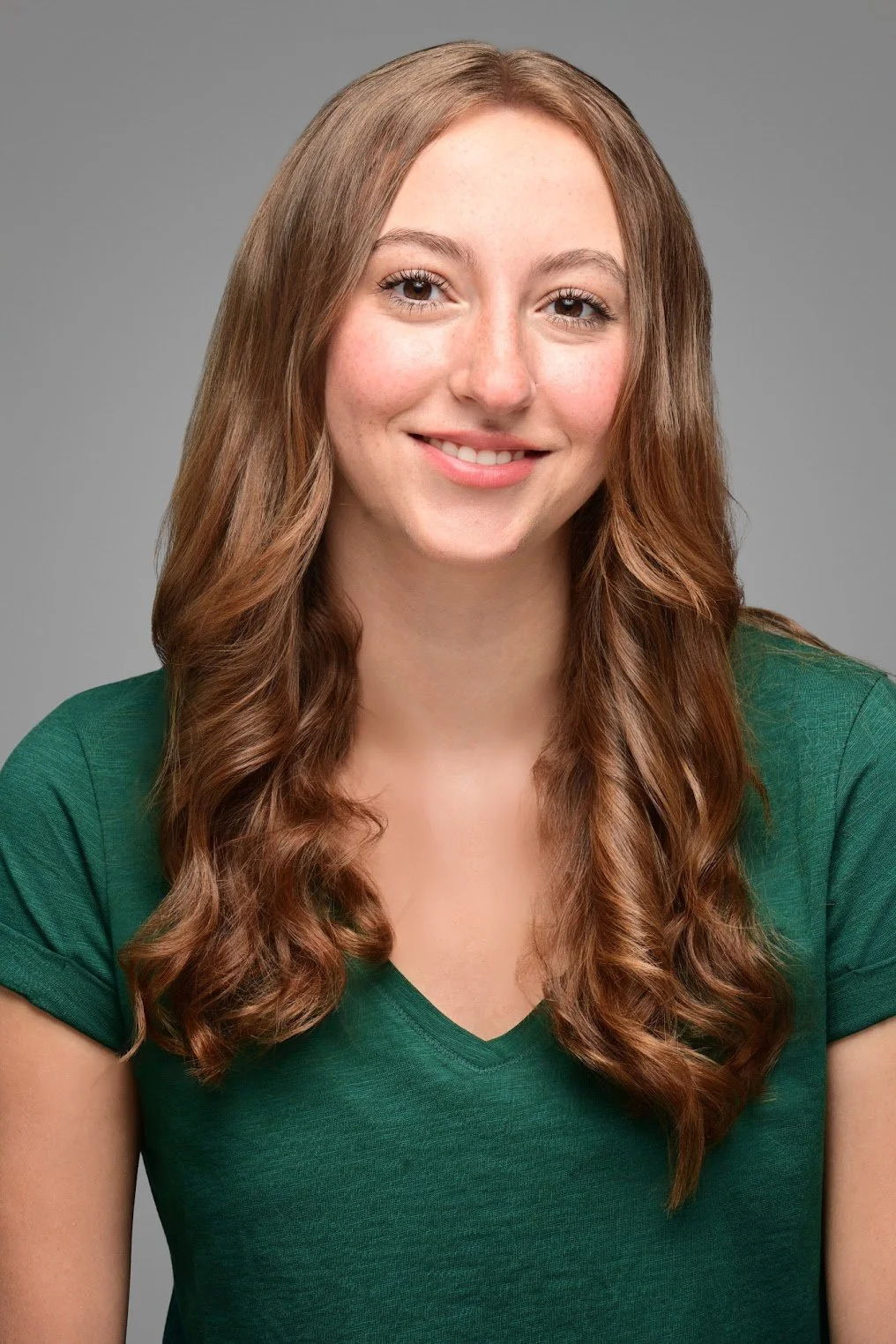 Portrait of Jiana Casazza, a young actress with long, wavy brown hair, smiling, wearing a green T-shirt, against a neutral gray background.