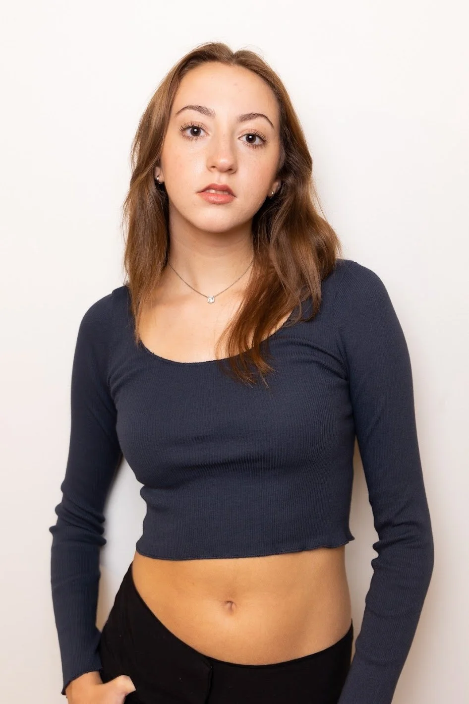 Photo of Jiana Casazza, a young actress with light skin and long wavy brown hair, wearing a navy blue long-sleeve crop top, black pants, and a small necklace, standing against a plain white background.