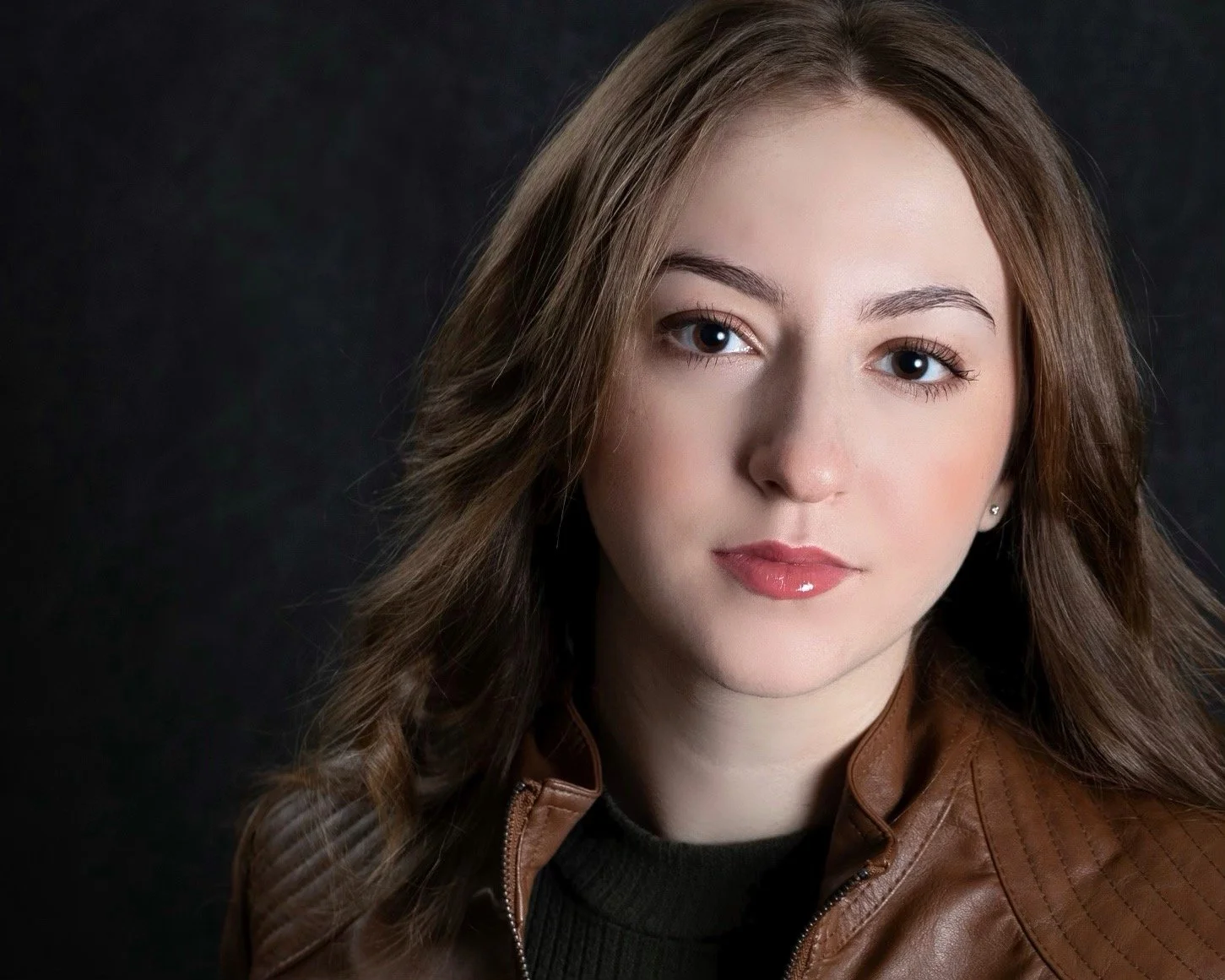 Landscape photo of actress Jiana Casazza. She is a young woman with long, wavy auburn hair, wearing a dark green ribbed sweater and brown leather jacket, against a dark background.