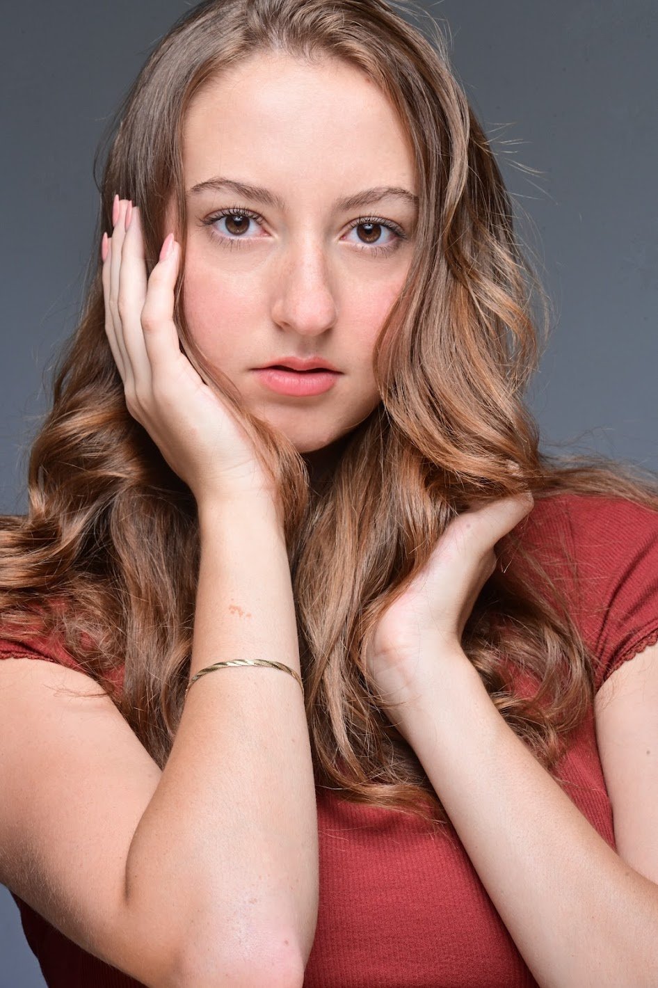 Close-up portrait of a Jiana Casazza, a young actress with long, wavy auburn hair, fair skin, and amber eyes, wearing a red top, touching her face gently, against a gray background.