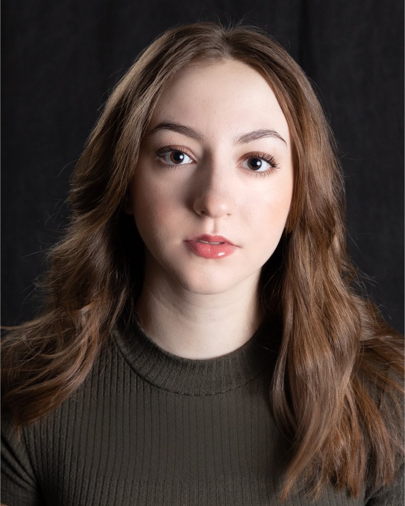 Photo of actress Jiana Casazza. She is a young woman with long, wavy auburn hair, wearing a dark green ribbed sweater and brown leather jacket, looking directly at the camera with a neutral expression against a dark background.