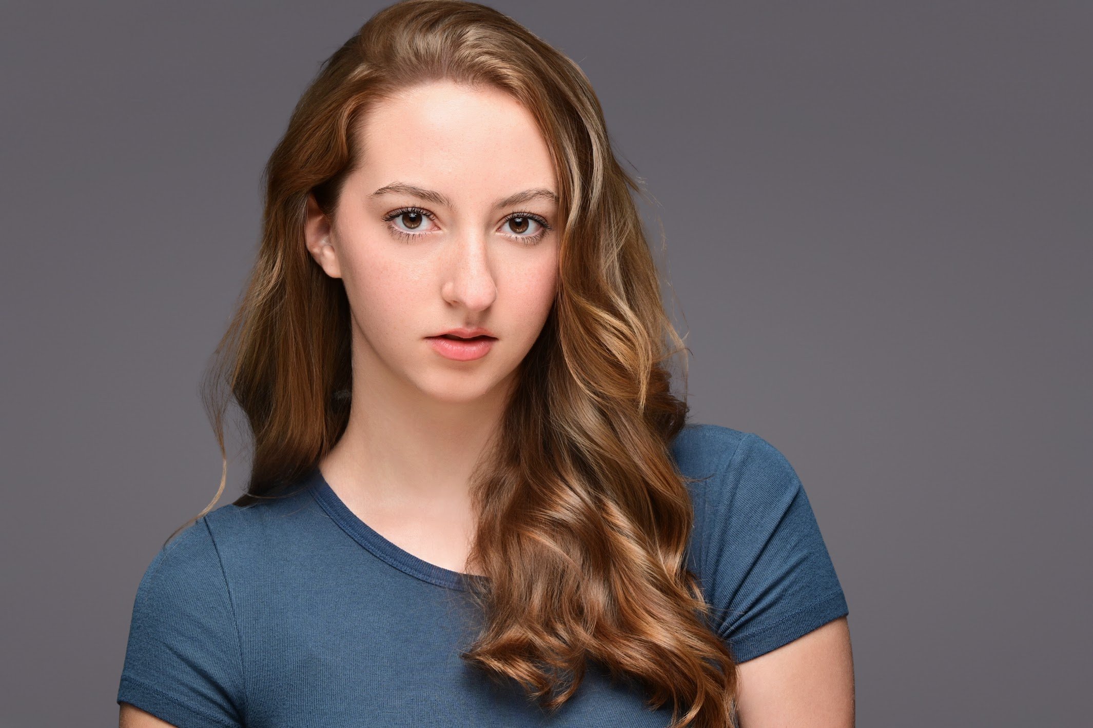 A young woman with wavy auburn hair and fair skin wearing a blue t-shirt against a gray background.