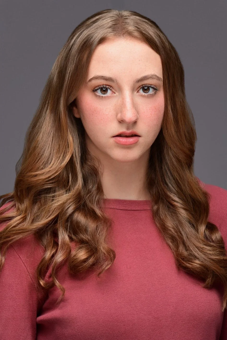 Portrait of Jiana Casazza, a young actress with long, wavy brown hair, fair skin, and wearing a pink top, against a gray background.
