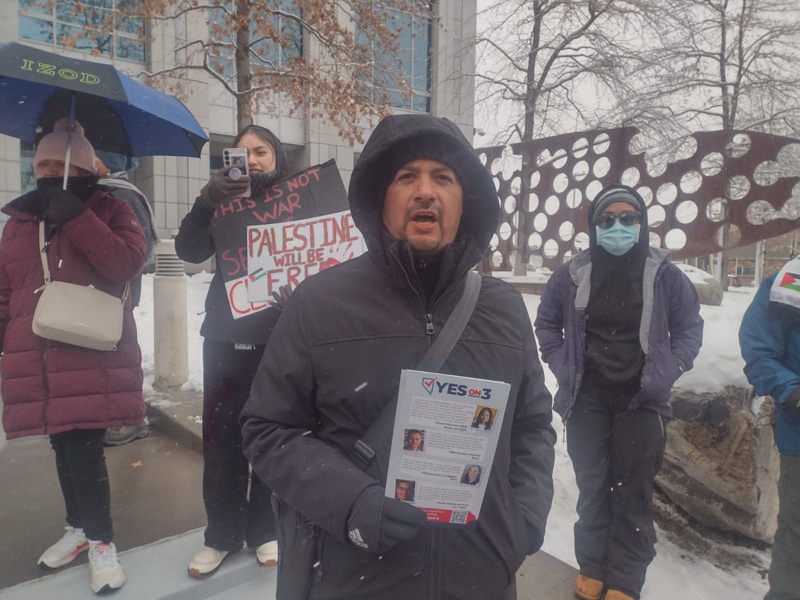 A group of people protesting outdoors in winter, holding signs and flyers, some wearing masks and winter clothing, with snow on the ground and leafless trees in the background.