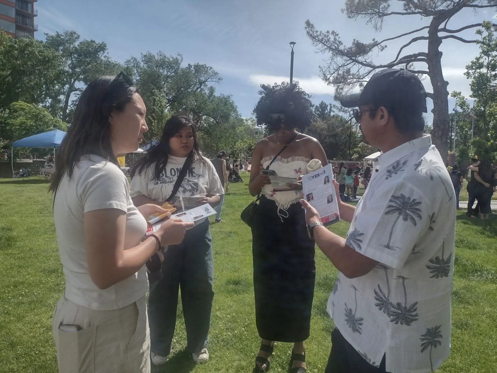 Four people standing outdoors on a grassy area, three women and one man, engaged in conversation, holding flyers or pamphlets. The background shows trees, a blue canopy, and other people in the distance.