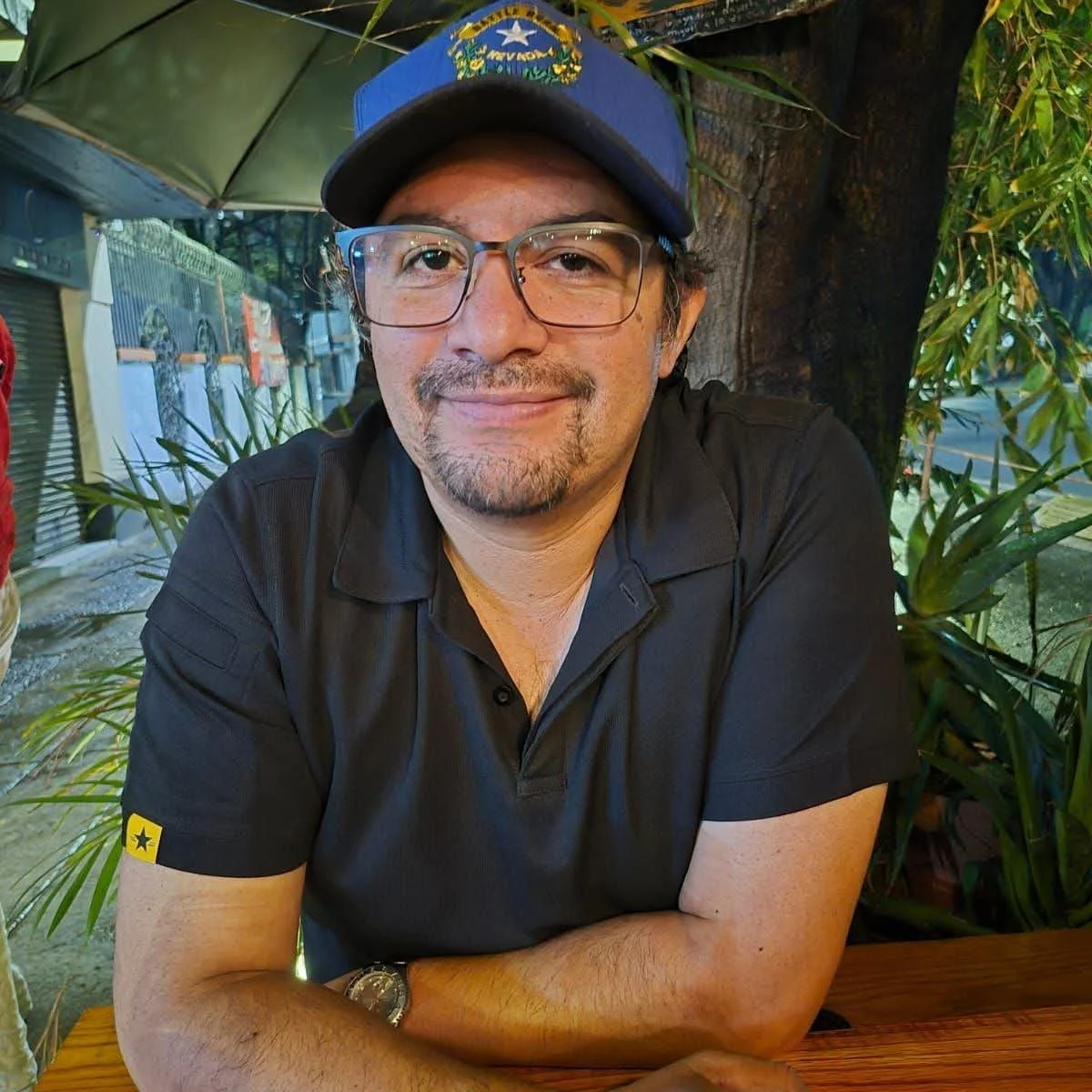A man with glasses and a goatee smiling, wearing a navy blue cap, a black polo shirt, and a watch, sitting at a wooden table outside, surrounded by green plants and trees.