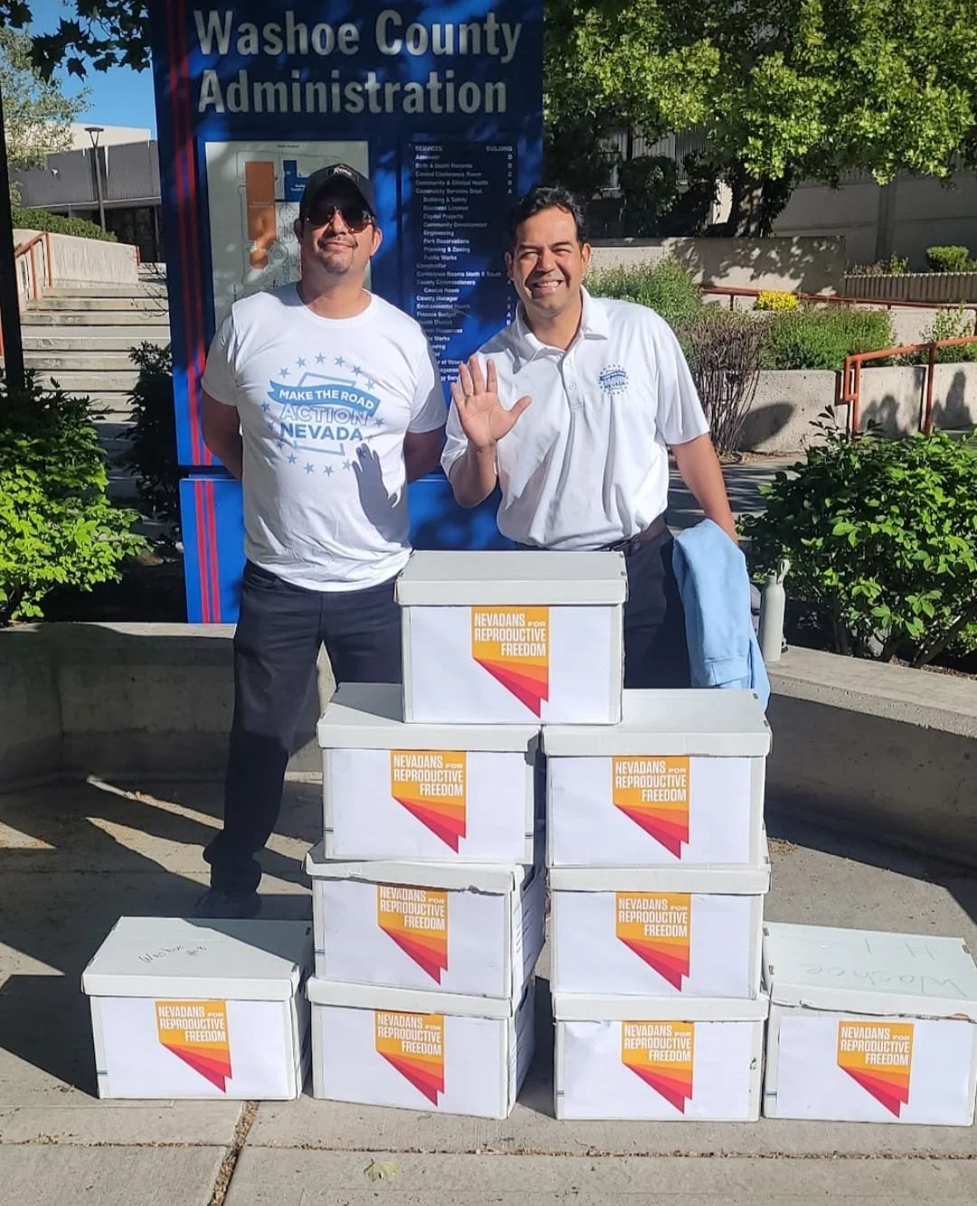 Two men standing outdoors in front of a large blue sign that says 'Washoe County Administration'. They are smiling and waving at the camera. There are several boxes with labels that read 'Nevadans for Reproductive Freedom' in front of them. The man on the left is wearing sunglasses and a white t-shirt with a blue and white design, while the man on the right is dressed in a white button-up shirt and holding a light blue jacket.