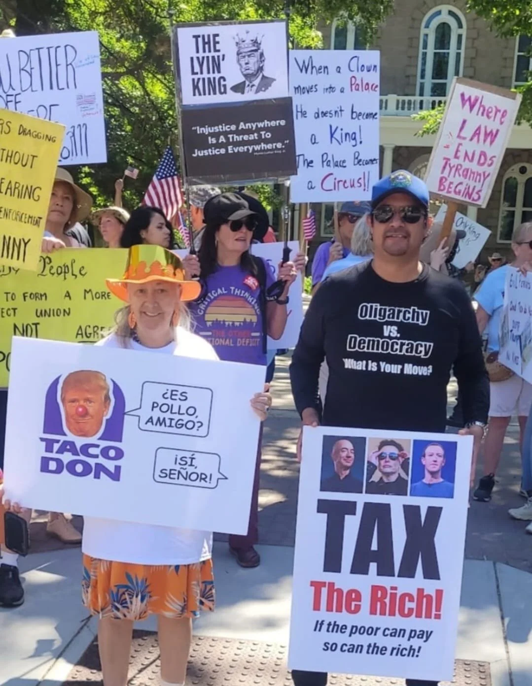 Group of protesters holding signs and banners during a demonstration advocating for political issues. Signs include slogans like 'Oligarchy vs. Democracy,' 'Tax the Rich,' and 'Injustice Anywhere Is A Threat To Justice Everywhere,' with some people wearing patriotic clothing and accessories.