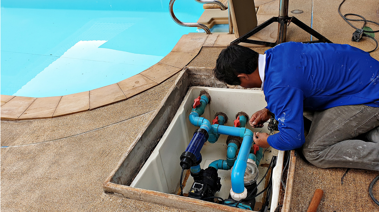 A man in a blue shirt kneeling down and working on pool equipment, installing or repairing plumbing near a swimming pool.