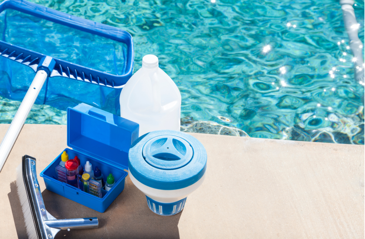Swimming pool cleaning supplies including a pool skimmer net, a blue toolbox with bottles of chemicals, a large white jug, a pool chlorine dispenser, and a pool skimmer basket, set on the pool deck by the water.