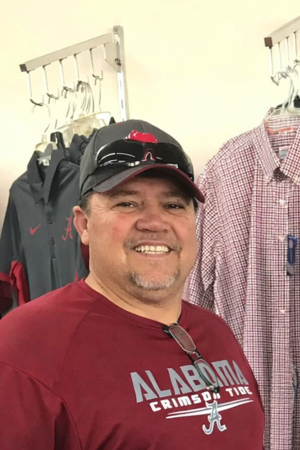 A smiling man wearing a black and red cap, a red Alabama Crimson Tide t-shirt, and sunglasses on his cap, standing in front of a rack with hanging clothes and a wall with patterned shirts.