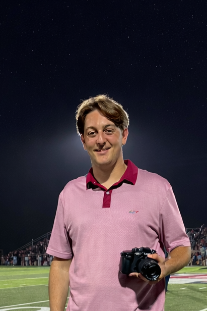 A young man in a pink polo shirt holding a camera, standing on a football field at night with a starry sky in the background.