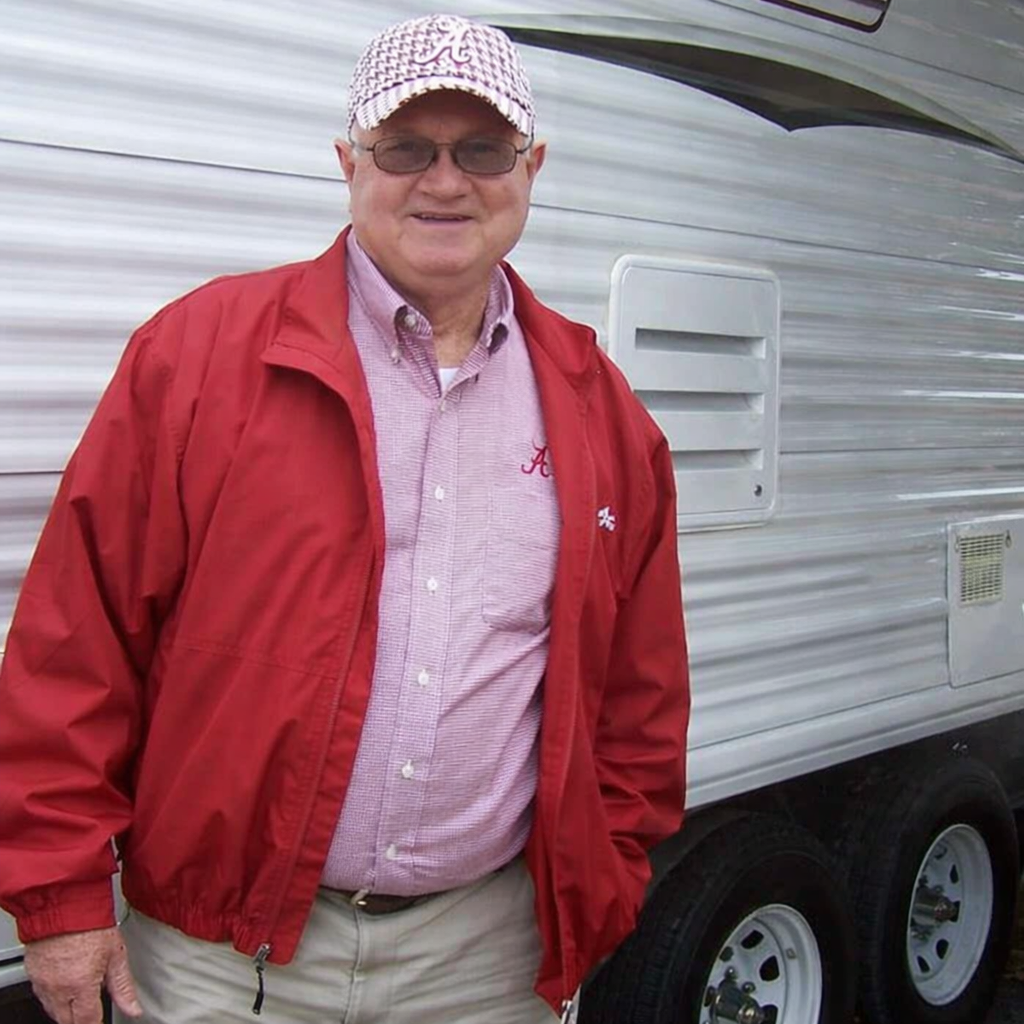 A smiling man wearing glasses, a patterned cap, a red jacket, a pink checkered shirt, and beige pants standing next to a white trailer with vents and black tires.