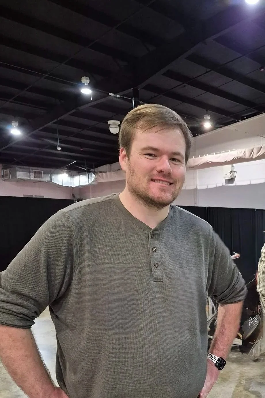A man with light brown hair and a beard, wearing a gray long-sleeve henley shirt, smiling with his hands on his hips in an indoor setting with black curtains and ceiling lights.