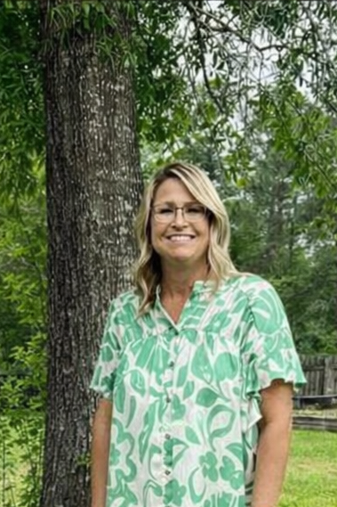 A woman with blonde hair and glasses smiling outdoors, wearing a green and white patterned shirt, standing next to a tree.