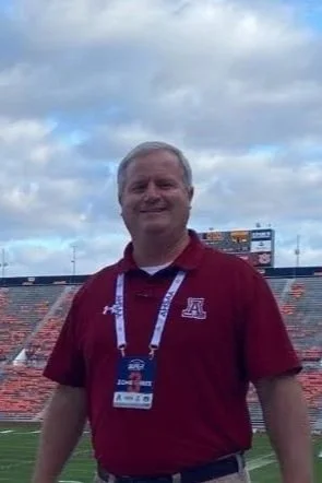 A man in a maroon polo shirt with an ID badge, standing in a sports stadium, with empty stadium seats and a cloudy sky in the background.